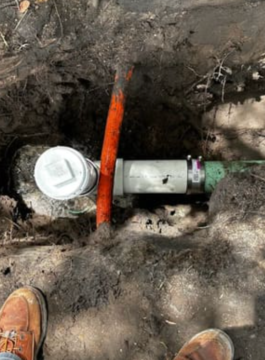 An excavated trench showing a newly installed white PVC pipe with a cleanout cap connected to an older green pipe.