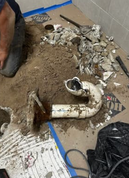 A person kneels by a section of white PVC plumbing pipe unearthed from broken concrete on a floor.