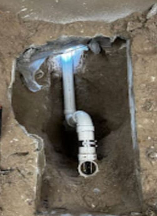 A section of white PVC plumbing pipe with a flexible coupler visible inside a hole dug into dirt beneath concrete.