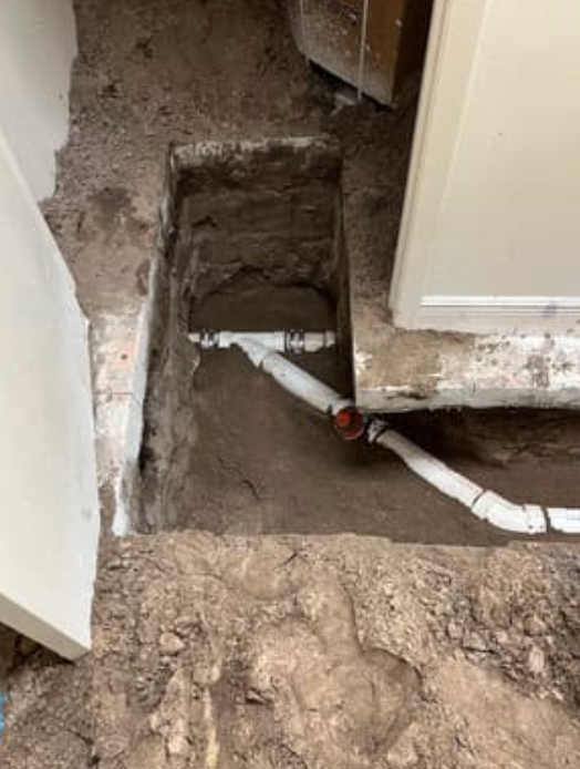 A rectangular hole in the ground reveals a section of white PVC plumbing pipe with a T-junction connector.