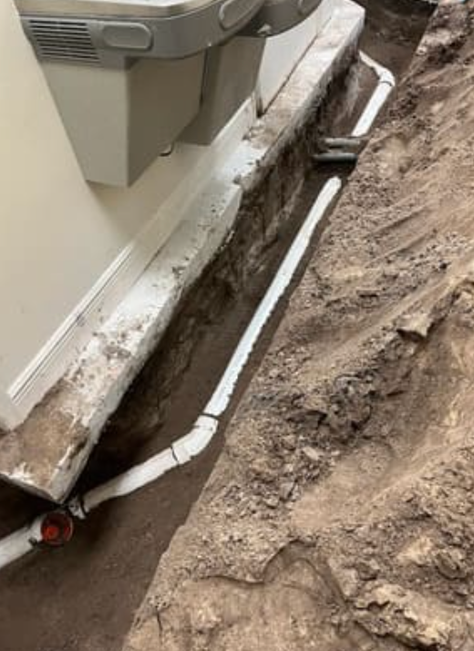 A trench alongside a building wall contains white PVC piping connecting to the structure's drainage system.