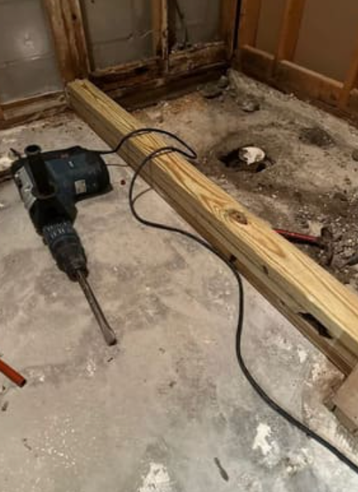 A heavy-duty drill sits on a concrete floor next to a wooden beam and a hole in the ground during a renovation project.