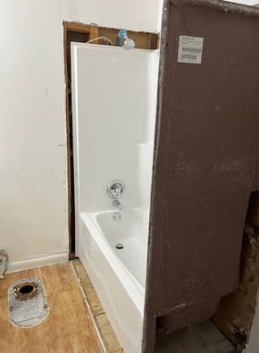 A partially installed white bathtub and shower surround in a room undergoing construction with exposed wall studs.