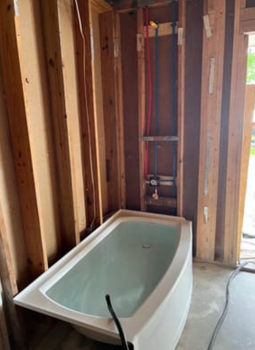 A bathtub installed in an unfinished bathroom with exposed wall studs and red and blue plumbing pipes in the wall.