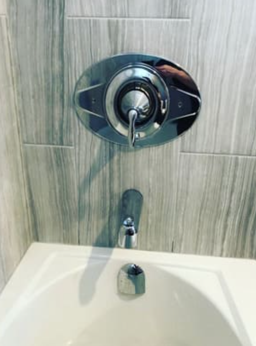 Chrome bathtub faucet and shower handle mounted on a light gray tiled wall above a white tub basin.