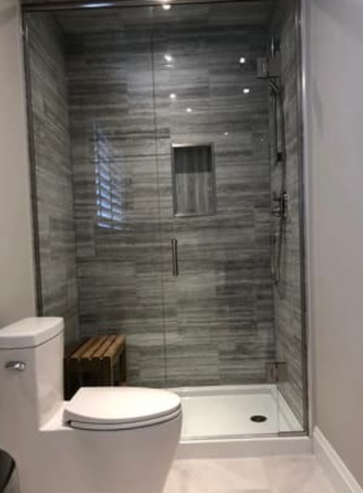 A white toilet next to a glass-enclosed shower with gray tiled walls and a small wooden stool inside.