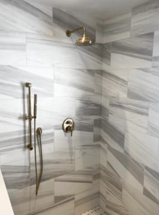 A shower stall featuring white and grey marble-style tiled walls with brushed bronze fixtures and a handheld spray.