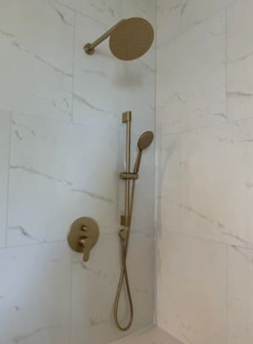 A gold-toned shower fixture with a rainfall head and a handheld sprayer on a slide bar against white marble-patterned tiles.