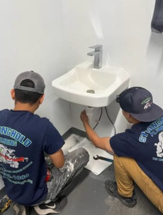 Two plumbers in company uniforms kneeling and working on the plumbing of a wall-mounted sink in a plain, white room.