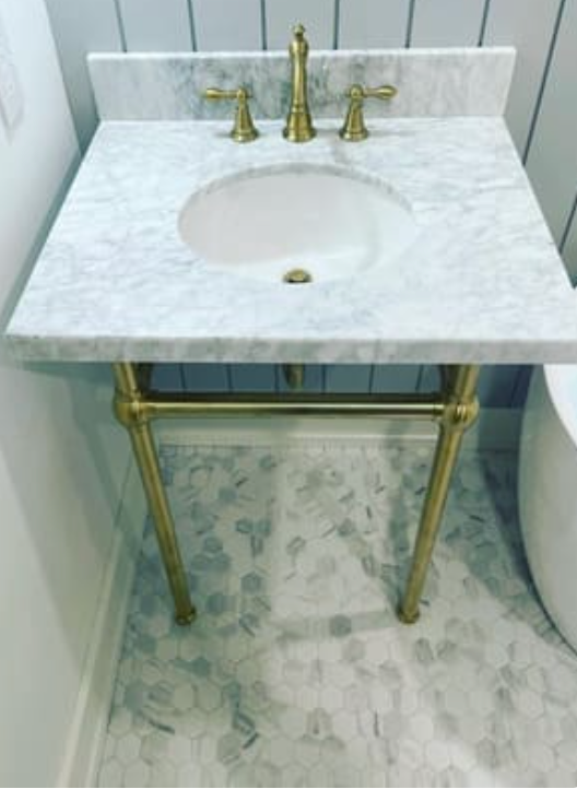 A white marble-topped bathroom sink with brass faucet and legs against a grey paneled wall and mosaic tile floor.