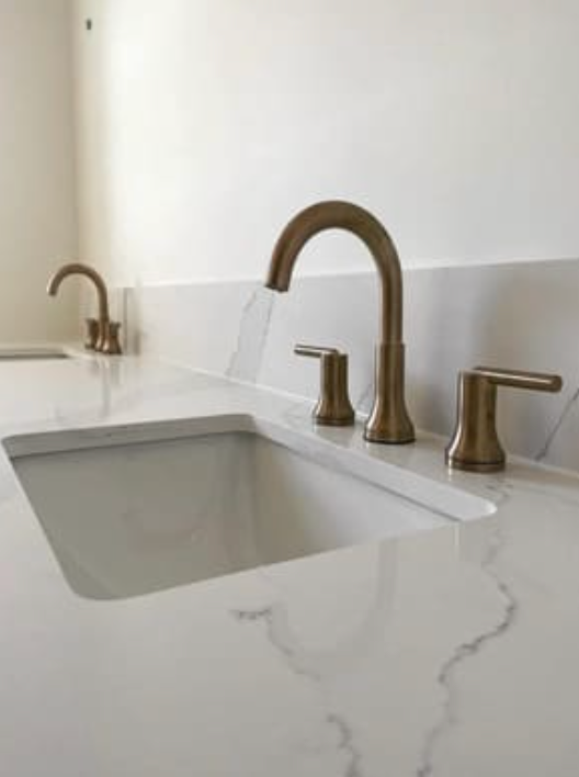 Two brass-toned bathroom faucets with lever handles installed on a white marble countertop with integrated sinks.