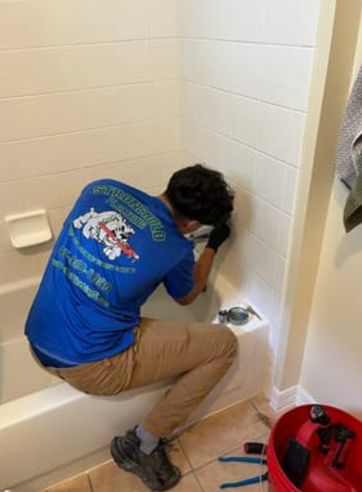 A plumber wearing a blue uniform works on a bathtub faucet in a tiled bathroom.