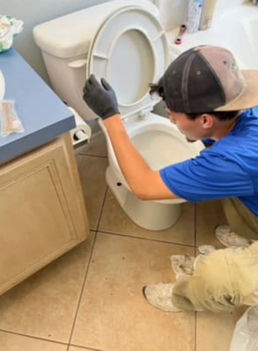 A worker in a blue shirt and cap wearing gloves inspecting a white toilet in a tiled bathroom.