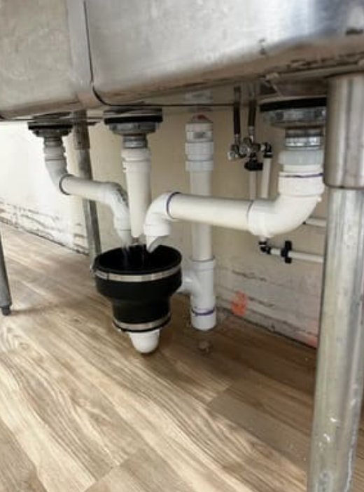 A stainless steel three-compartment sink drain system with white PVC pipes draining into a central black rubber funnel.