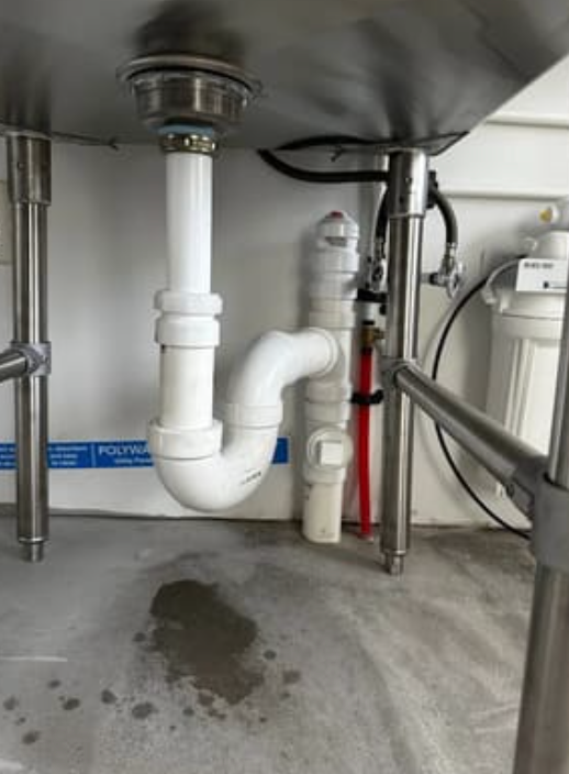 A white P-trap drain assembly connected to a stainless steel sink, with a small water puddle on the concrete floor below.
