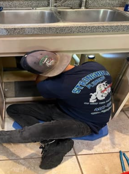 A plumber wearing a branded navy blue shirt and baseball cap works under a kitchen sink while kneeling on a blue pad.