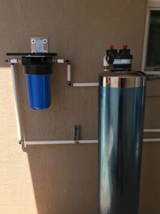 A blue water filter housing is mounted on a wall next to a tall, blue metallic water treatment tank connected by piping.