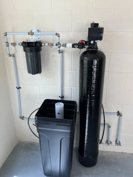 A home water filtration and softening system mounted against a light-colored cinder block wall.