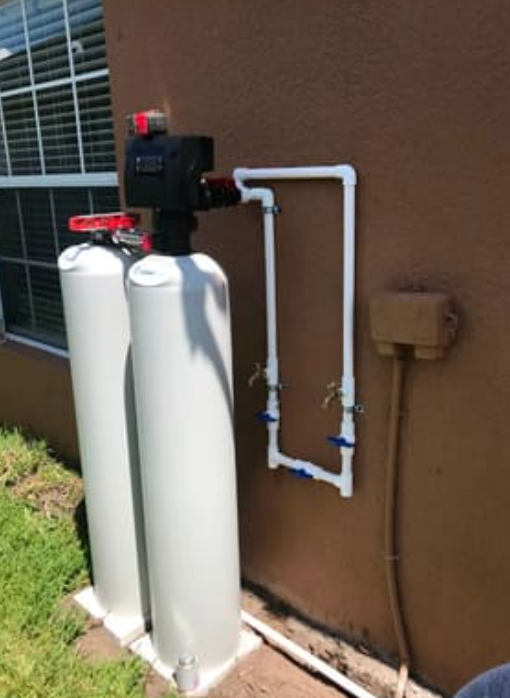 A water softening system with two white tanks and a control head mounted on an exterior wall with PVC bypass plumbing.