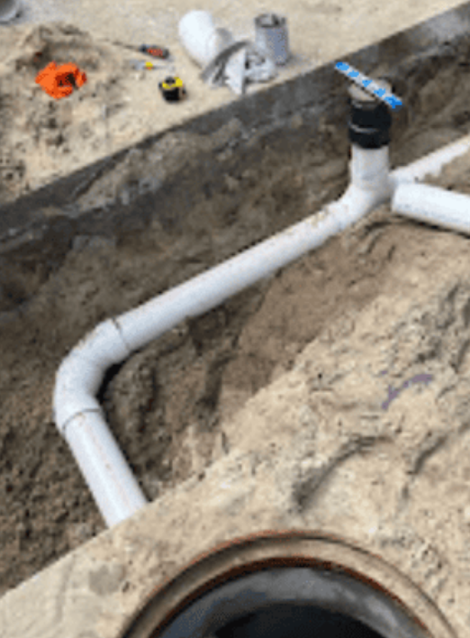 White PVC drainage pipes connected by a T-joint and elbow, resting in a dirt trench next to a circular concrete opening.