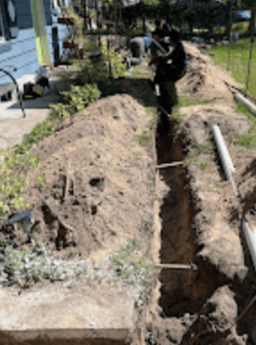 Two people work in a long, narrow trench dug into the dirt along the side of a house.