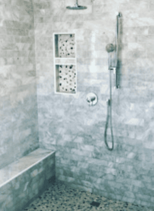 A walk-in shower with stone tile walls, a pebble floor, a bench, a rainfall showerhead, and a built-in storage niche.
