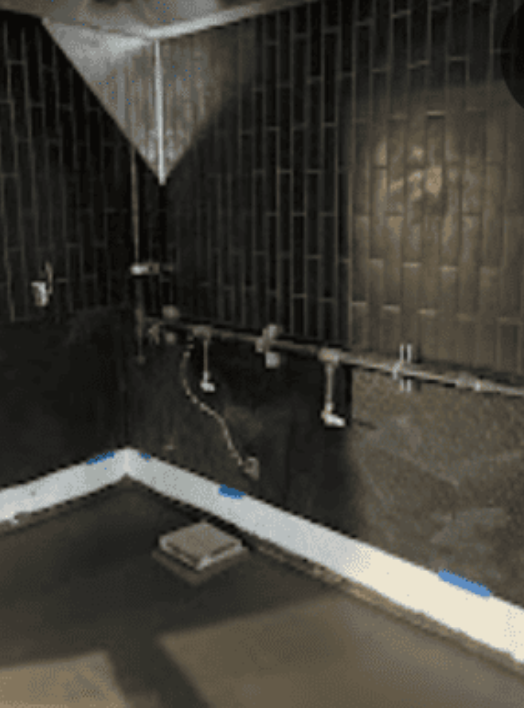 A room under construction featuring dark, vertical-tiled walls, exposed silver plumbing pipes, and an unfinished floor.