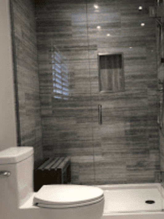 A bathroom featuring a white toilet next to a glass-enclosed shower with grey horizontal tiles and a recessed wall niche.