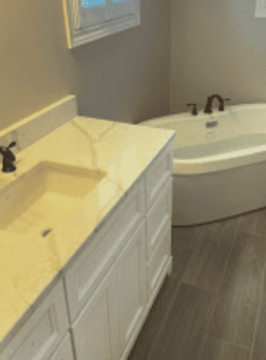 A white bathroom vanity with a marbled countertop sits beside a freestanding white soaking tub on gray tiled floors.