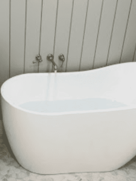 A modern white freestanding bathtub being filled by a wall-mounted faucet against a light gray vertical-paneled wall.