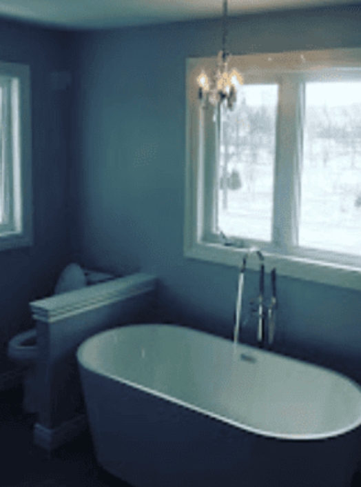 A freestanding white bathtub with a floor-mounted faucet sits before a large window in a gray-walled bathroom.