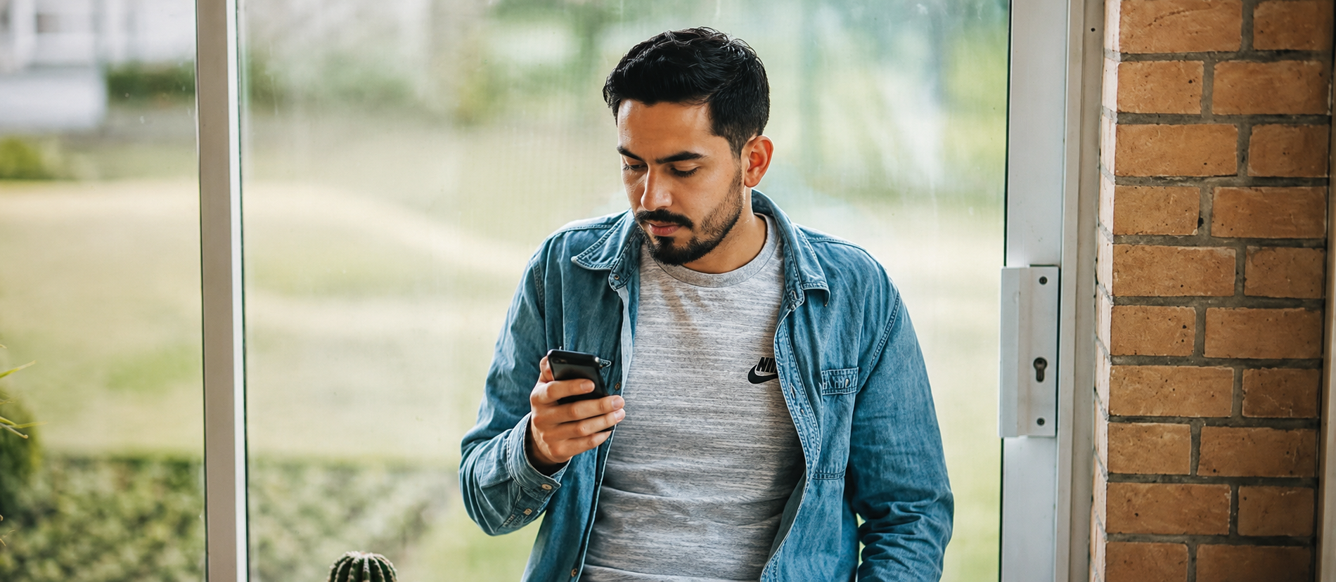 Man in a denim shirt reading his phone beside a window and small potted plant