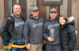 Four people in dark hoodies posing outside, holding a 2018 Torch Awards trophy.