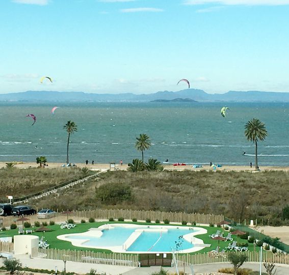 Beach scene with kitesurfers, pool, palm trees, and mountains in the distance.