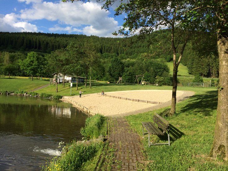 Lakeside sandy beach area with a brick path, bench, and wooded hillside under a blue sky.