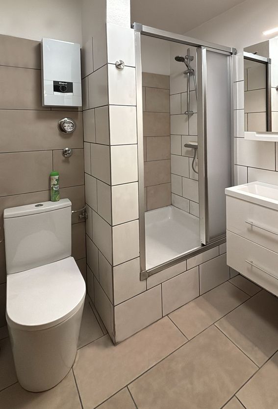 Small modern bathroom with a toilet, shower, and vanity. Beige and white tiles cover the walls and floor.