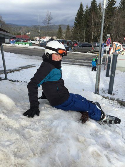 Person in ski gear sitting on snow near a parking lot, wearing helmet and goggles.
