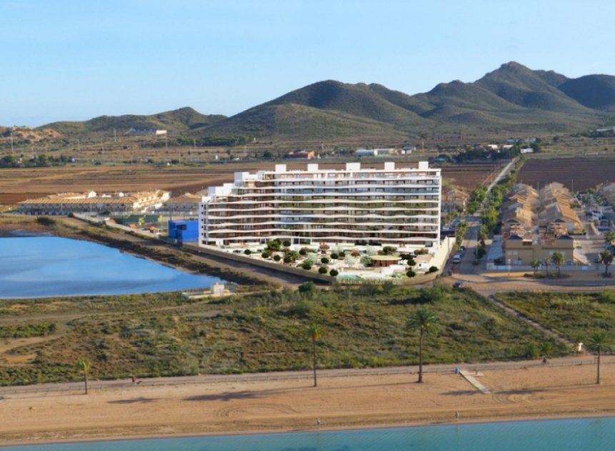 Modern white building with balconies beside a lake and mountains under a sunny sky.