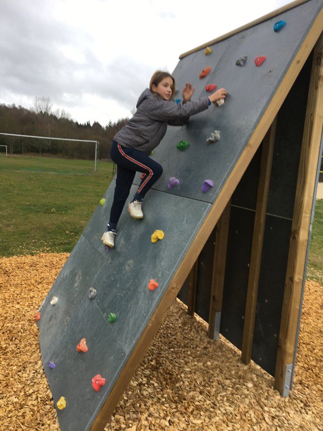 A person climbs an angled climbing wall with colorful holds on wood chips; a field and trees are in the background.