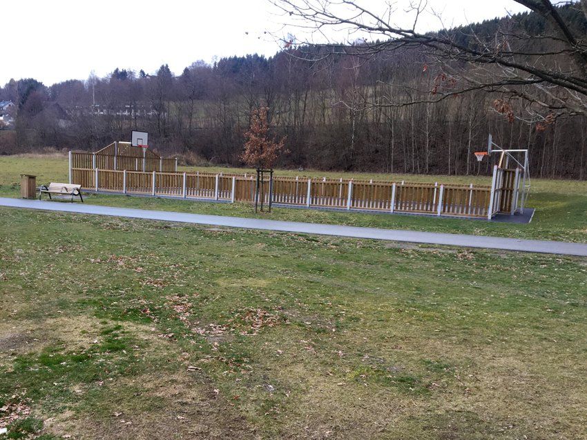 A basketball court with wooden fencing, set in a grassy park, a path in front, and a treeline in the background.
