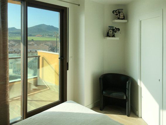 Bedroom with sliding glass door, black armchair, white shelves with decorations, and a view of mountains.