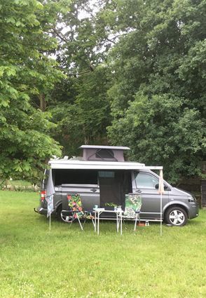 Gray campervan parked on grass with awning extended and two chairs. Trees in background.