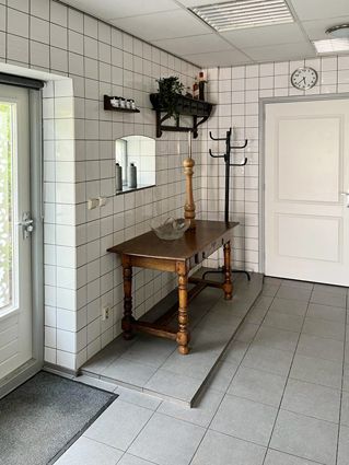 Interior hallway with tiled walls and floor. A table sits on a small ramp. Door and coat rack visible.