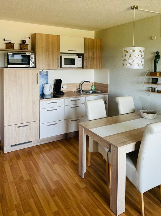 Small kitchen with cabinets, microwave, and dining table with chairs. Wooden floor.
