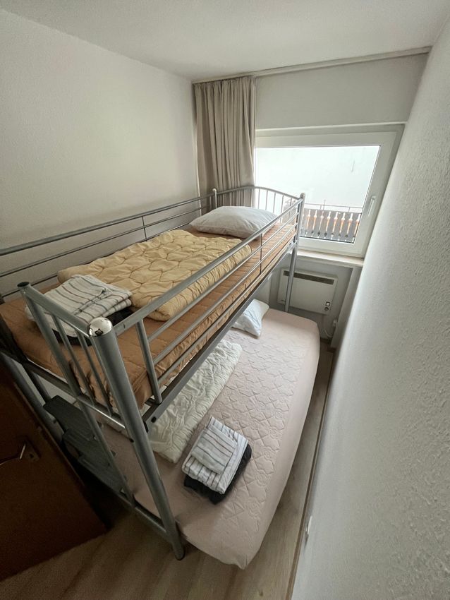 Bunk beds in a small room with a window. The top bed has a yellow blanket, the bottom has a pink mattress.