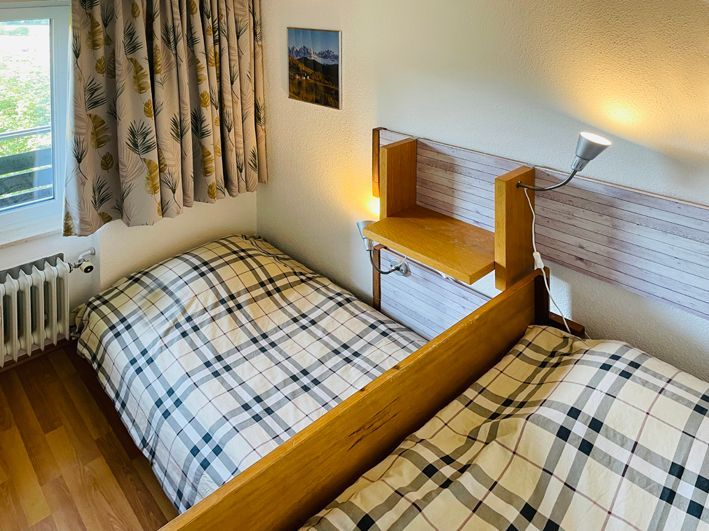 Two twin beds with plaid bedding in a room with a small shelf, window, and patterned curtains.