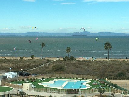 Beach scene with kitesurfers, palm trees, and a swimming pool under a blue sky.