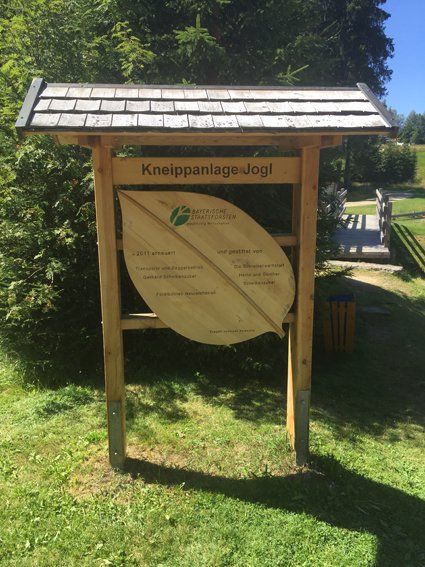 Wooden sign for Kneippanlage-Jogl, in green grass, with bridge and forest in the background.