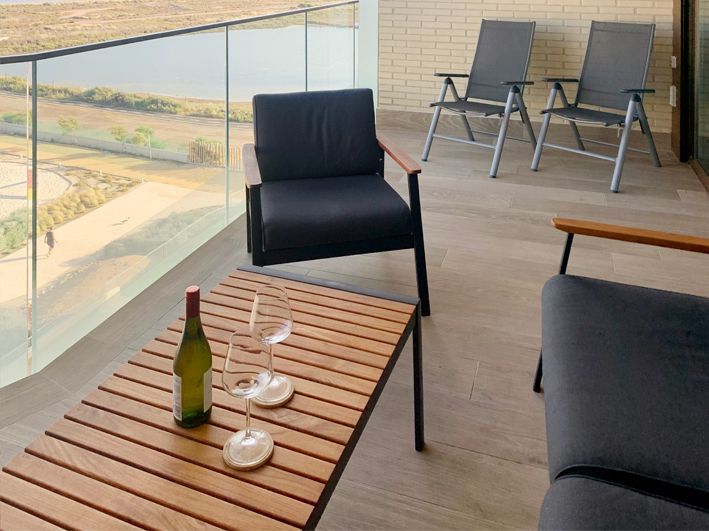 Balcony with modern furniture: table with wine and glasses, chairs, and glass railing overlooking a landscape.