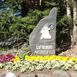 Stone sign for Luftkurort Niedersfeld, with a goat emblem, surrounded by flowers.